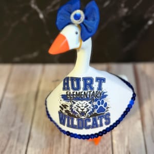 May include: A white decorative goose figurine wearing a blue fabric bow with a central rhinestone accent on its head. The goose is dressed in a white cape featuring the text Hurt Elementary Wildcats, a graphic of a wildcat face, and a blue paw print.