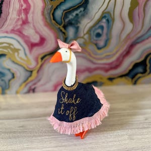 May include: A decorative goose figurine with a white body, orange beak and feet, and a pink bow. It wears a navy blue cape with pink fringe and gold embroidered text that reads "Shake it off."
