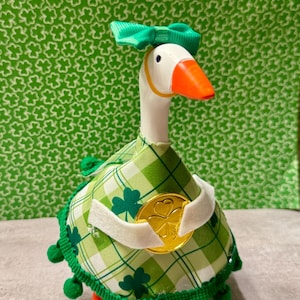 May include: A St. Patrick's Day themed goose ornament. The goose has a white head, orange beak, and is wearing a green plaid outfit with shamrocks and a gold coin. A green bow is on its head.