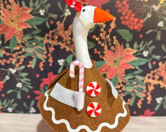 May include: A white goose figurine with an orange beak and feet, wearing a gingerbread-themed costume. The costume includes a brown felt dress with white trim, candy buttons, and a candy cane. A red and white hat is on its head. The background features a floral pattern.