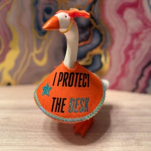 Może przedstawiać: Figurka gęsi z pomarańczowym filcowym kołnierzem z napisem "I PROTECT THE DESK". Kołnierz jest ozdobiony turkusową gwiazdą i obszyciem z koralików. Gęś ma pomarańczowy dziób i stopy oraz mały pomarańczowy element na głowie.