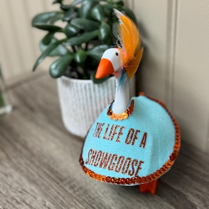 Peut inclure: Figurine d&eacute;corative de canard avec une t&ecirc;te blanche, un bec et des pieds oranges, et un corps bleu clair. Le corps est orn&eacute; de paillettes oranges et porte l'inscription "THE LIFE OF A SHOWGOOSE". Le canard porte une plume orange. Une plante dans un pot blanc est en arri&egrave;re-plan.