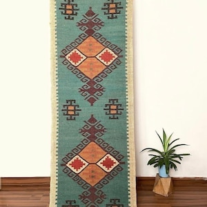May include: A long, rectangular runner rug featuring a teal green base with a repeating geometric pattern in dark brown, orange, and red. The rug has a light beige border and includes the text MAMTAEXPORTS printed at the bottom center.