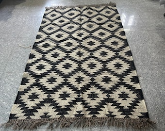 May include: A rectangular rug with a geometric pattern in black and beige. The design features repeating diamond shapes. The rug has a fringed edge in a brown color. The rug is made of woven material.