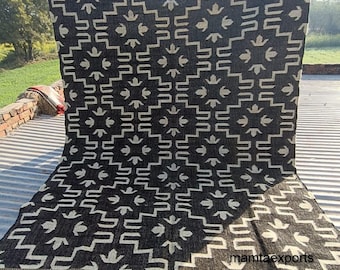 May include: A large, rectangular black and beige patterned rug. The rug features a repeating geometric design with a fringe along the bottom edge. The text "mamtaexports" is visible on the rug. The rug is displayed outdoors.
