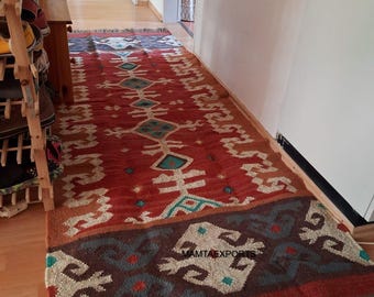 May include: A long, rectangular rug with a red background and geometric patterns in teal, beige, and brown. The rug has a border with a repeating design and fringe at the end. The text "MAMTAEXPORTS" is visible on the rug.