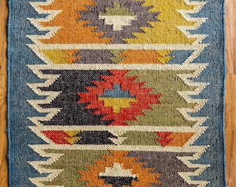 May include: Rectangular blue rug with a geometric pattern in shades of orange, yellow, red, and gray. The rug has a fringed edge and a woven texture. The text "mamtaexports" is visible near the bottom.