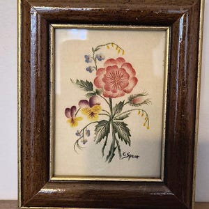 Puede incluir: Pintura floral en acuarela enmarcada. La obra presenta un ramo de flores rojas, moradas, amarillas y azules con hojas verdes. El marco es marrón oscuro con un borde interior dorado. La firma del artista, "C. Spear", es visible.
