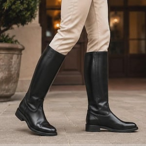 Puede incluir: Un par de botas negras hasta la rodilla con acabado liso, punta redondeada y tacón bajo de bloque. Las botas presentan una caña estructurada que llega justo debajo de la rodilla, con detalles de costuras visibles en la zona del tobillo.