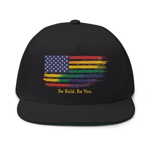 Peut inclure: Casquette noire à visière plate avec un drapeau américain délavé aux couleurs de l'arc-en-ciel. Le drapeau présente des étoiles et des rayures, avec le texte "Be Bold. Be You." en jaune. La casquette a un dessous vert.