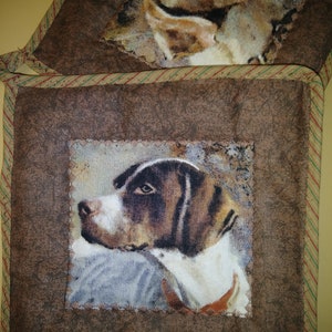 May include: Two brown fabric pot holders with a brown and white dog illustration on each. The dog's head is facing to the left. The pot holders have a brown, green, and red striped border.