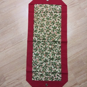 May include: A red and cream table runner with a green holly and ribbon pattern. The runner has a red border and a button on each end.