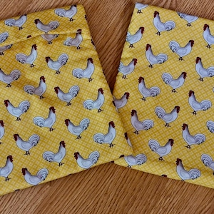 May include: Two square fabric pot holders featuring a repeating pattern of white roosters with red combs on a yellow background with a subtle white grid design. The items are flat, quilted, and display a consistent print of poultry across the surface.