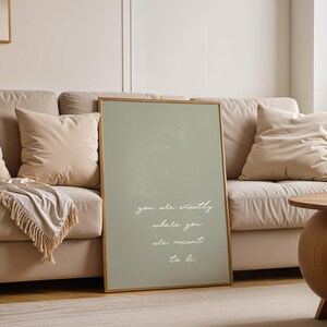 Puede incluir: Un estampado enmarcado con un fondo verde salvia y un marco de madera. El estampado presenta la cita "you are exactly where you are meant to be" en escritura blanca. El estampado est&aacute; apoyado contra un sof&aacute; beige con almohadas.