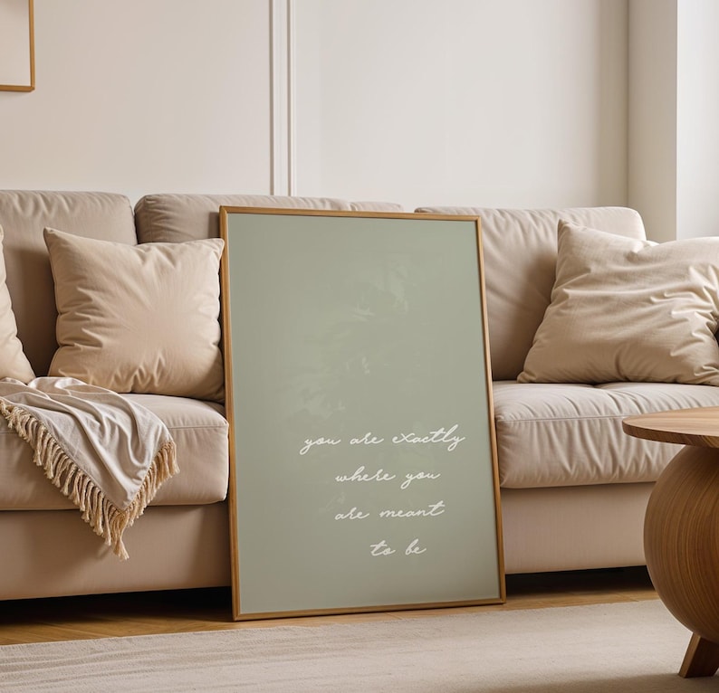 Puede incluir: Un estampado enmarcado con un fondo verde salvia y un marco de madera. El estampado presenta la cita "you are exactly where you are meant to be" en escritura blanca. El estampado est&aacute; apoyado contra un sof&aacute; beige con almohadas.