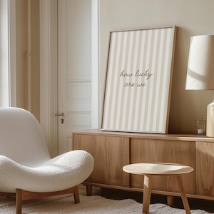 May include: A framed print with a beige and white striped pattern and the text "how lucky are we." The print is displayed on a wooden cabinet in a room with a white chair and a small wooden table.