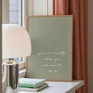 Puede incluir: Un cuadro enmarcado con un fondo verde salvia y el texto "you are exactly where you are meant to be" en escritura blanca. El marco es de madera clara, y el cuadro descansa sobre una superficie blanca con libros y una l&aacute;mpara plateada.