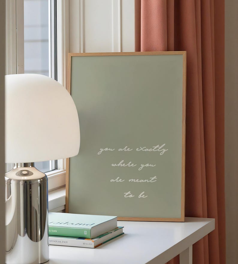 Puede incluir: Un cuadro enmarcado con un fondo verde salvia y el texto "you are exactly where you are meant to be" en escritura blanca. El marco es de madera clara, y el cuadro descansa sobre una superficie blanca con libros y una l&aacute;mpara plateada.