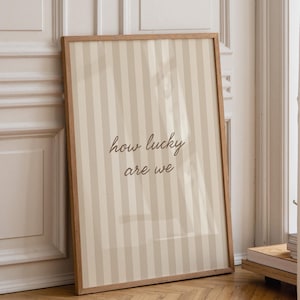 May include: A framed print with a light brown wooden frame. The print features a striped pattern in beige and off-white, with the words "how lucky are we" in a handwritten font.