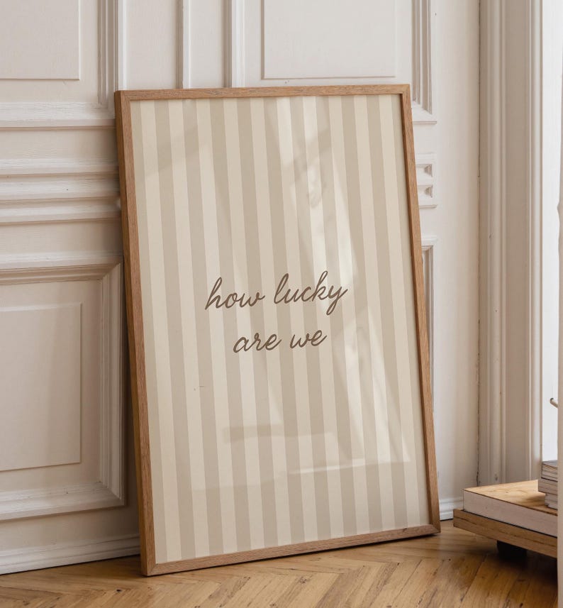 May include: A framed print with a light brown wooden frame. The print features a striped pattern in beige and off-white, with the words "how lucky are we" in a handwritten font.