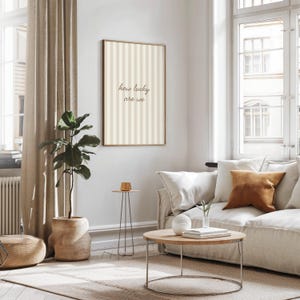 May include: A framed vertical print with a beige and white striped pattern and the text "how lucky are we" in a living room setting. The room includes a sofa, coffee table, side table, potted plant, and window with beige curtains.