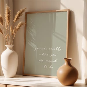 Puede incluir: Una impresi&oacute;n enmarcada con el texto "you are exactly where you are meant to be" en escritura blanca. La impresi&oacute;n tiene un fondo verde apagado y un marco de madera clara. Un jarr&oacute;n blanco con plantas secas y un jarr&oacute;n marr&oacute;n est&aacute;n en primer plano.