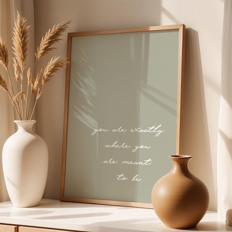 Puede incluir: Una impresi&oacute;n enmarcada con el texto "you are exactly where you are meant to be" en escritura blanca. La impresi&oacute;n tiene un fondo verde apagado y un marco de madera clara. Un jarr&oacute;n blanco con plantas secas y un jarr&oacute;n marr&oacute;n est&aacute;n en primer plano.