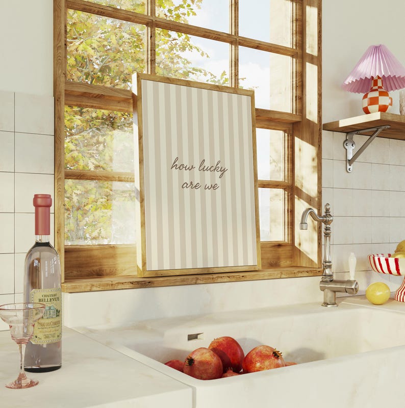 May include: A framed print with vertical beige and white stripes and the text "how lucky are we" sits on a wooden windowsill. A bottle of wine and a glass are on the counter. A sink with pomegranates is in the foreground.