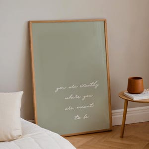 Puede incluir: Un cuadro enmarcado con un fondo verde salvia y la cita "you are exactly where you are meant to be" en escritura blanca. El marco es de madera clara. Una peque&ntilde;a maceta de cer&aacute;mica marr&oacute;n est&aacute; sobre una mesita auxiliar.