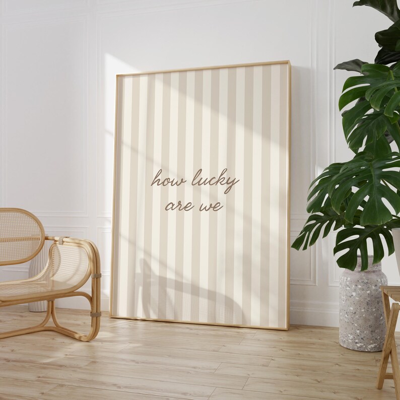 May include: A framed wall art print with a beige and cream striped pattern. The text "how lucky are we" is written in a cursive font. The artwork is displayed in a light-colored wooden frame.