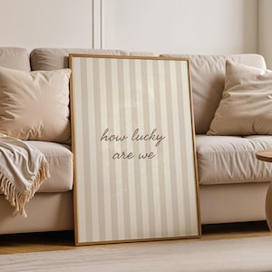 May include: A framed vertical print with a striped pattern in beige and off-white. The text "how lucky are we" is written in a cursive font. The print is leaning against a beige sofa with pillows and a throw blanket.