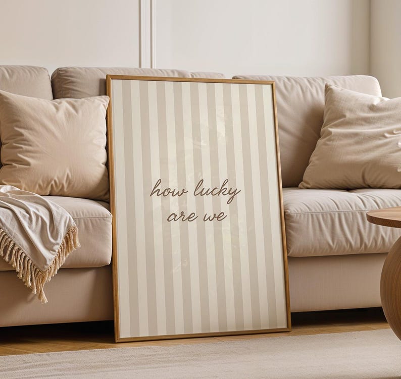 May include: A framed vertical print with a striped pattern in beige and off-white. The text "how lucky are we" is written in a cursive font. The print is leaning against a beige sofa with pillows and a throw blanket.