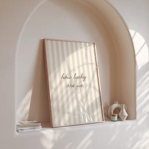 May include: A framed print with a beige and white striped pattern and the text "how lucky are we." The print is displayed on a shelf with decorative vases and a small candle. The frame is a light brown color.