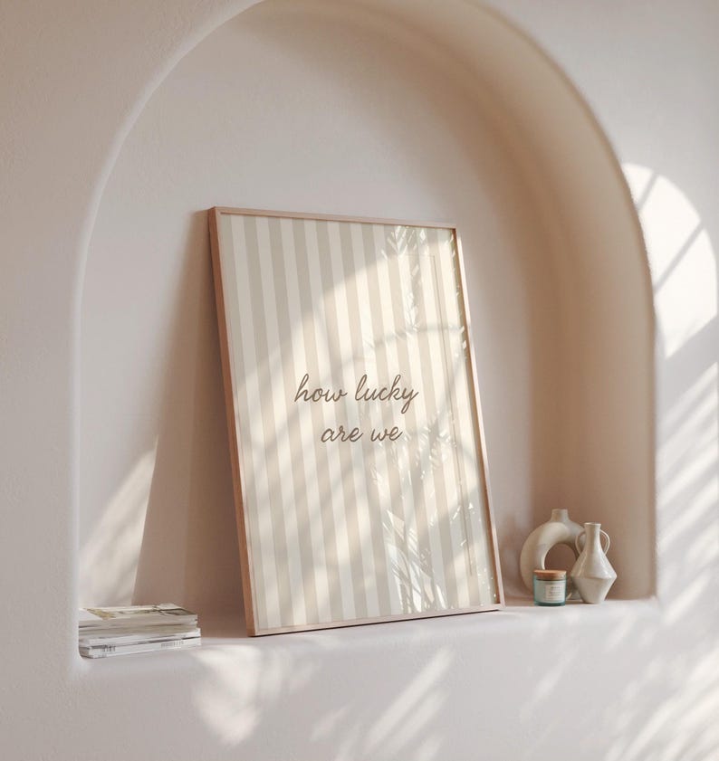May include: A framed print with a beige and white striped pattern and the text "how lucky are we." The print is displayed on a shelf with decorative vases and a small candle. The frame is a light brown color.