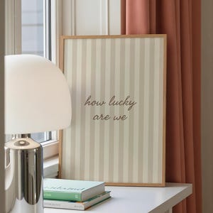 May include: A framed print with a striped background in beige and off-white, featuring the text "how lucky are we" in a cursive font. The print is displayed on a white surface, next to a lamp and books.