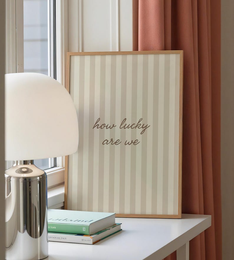 May include: A framed print with a striped background in beige and off-white, featuring the text "how lucky are we" in a cursive font. The print is displayed on a white surface, next to a lamp and books.
