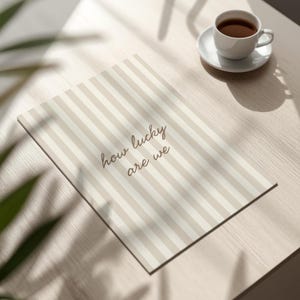 May include: A rectangular print with vertical beige and white stripes and the text "how lucky are we" in a cursive font. A white teacup and saucer with coffee sits on a light wood surface.