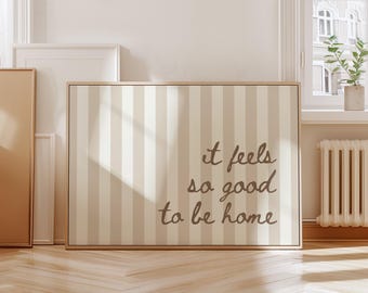 Pode incluir: Impressão em tela emoldurada com um padrão listrado em bege e branco sujo. O texto "it feels so good to be home" está escrito em itálico. A impressão é exibida em uma sala com uma janela e pisos de madeira.
