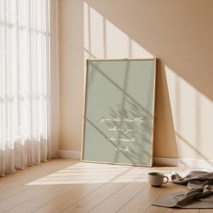 Puede incluir: Una impresi&oacute;n art&iacute;stica enmarcada con un fondo verde salvia y la cita "you are exactly where you are meant to be" en escritura blanca. La impresi&oacute;n se apoya en una pared en una habitaci&oacute;n con una ventana y una taza.