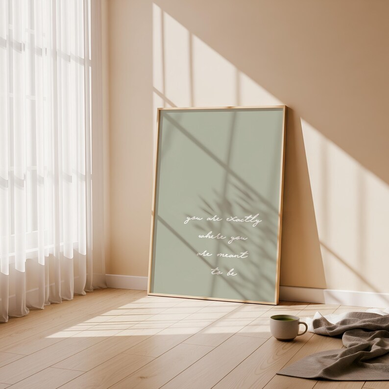 Puede incluir: Una impresi&oacute;n art&iacute;stica enmarcada con un fondo verde salvia y la cita "you are exactly where you are meant to be" en escritura blanca. La impresi&oacute;n se apoya en una pared en una habitaci&oacute;n con una ventana y una taza.
