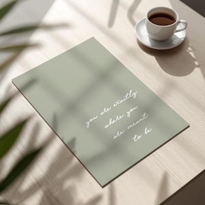 Puede incluir: Una impresi&oacute;n rectangular verde salvia con las palabras "you are exactly where you are meant to be" en escritura blanca. Una taza y platillo blancos con caf&eacute; sobre una superficie de madera clara.
