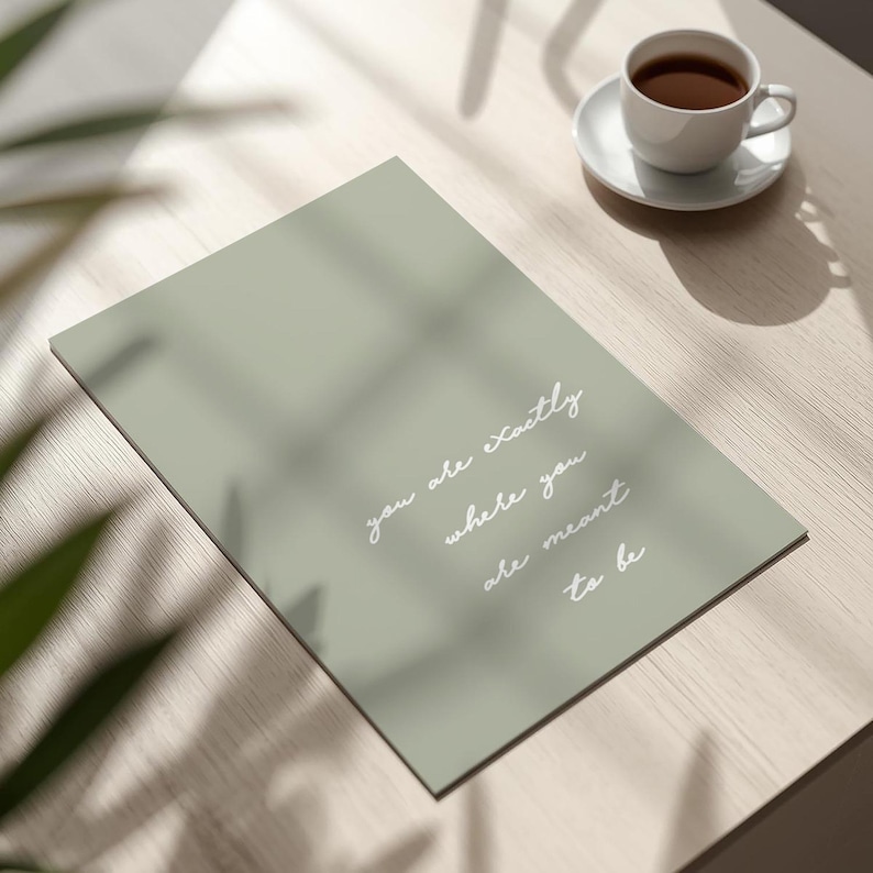 Puede incluir: Una impresi&oacute;n rectangular verde salvia con las palabras "you are exactly where you are meant to be" en escritura blanca. Una taza y platillo blancos con caf&eacute; sobre una superficie de madera clara.