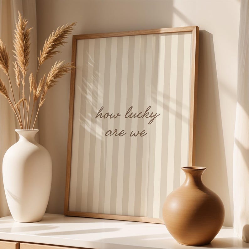 May include: A framed print with vertical beige and off-white stripes and the text "how lucky are we." A white vase with dried plants and a brown vase are on a white surface.
