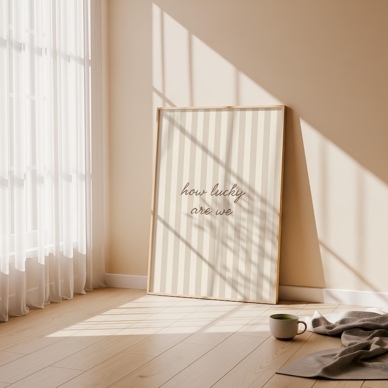 May include: A framed print with a vertical striped pattern in beige and off-white, leaning against a wall. The text "how lucky are we" is displayed in a cursive font. A mug and a folded blanket are on the wooden floor.