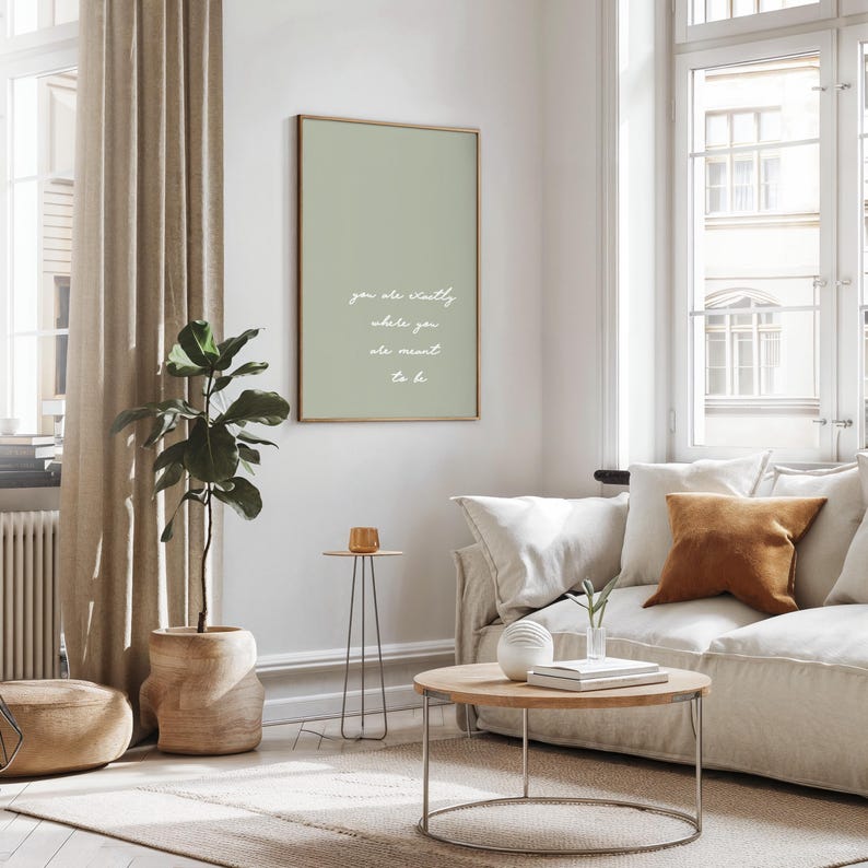 Puede incluir: Un sal&oacute;n moderno con un cuadro enmarcado verde salvia con la cita "you are exactly where you are meant to be". La habitaci&oacute;n incluye un sof&aacute; beige, una mesa de centro de madera y una planta en maceta. La decoraci&oacute;n es minimalista y contempor&aacute;nea.
