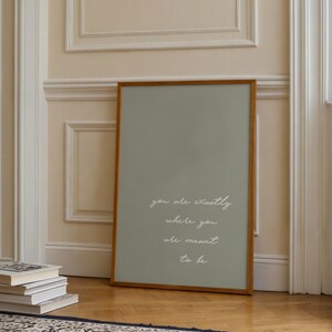 Puede incluir: Un estampado enmarcado con un fondo verde salvia y un marco de madera. El texto blanco en cursiva dice "you are exactly where you are meant to be". El estampado est&aacute; apoyado contra una pared junto a una pila de libros.