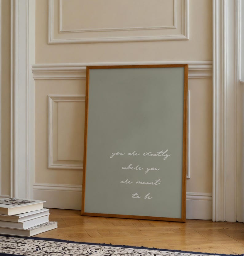Puede incluir: Un estampado enmarcado con un fondo verde salvia y un marco de madera. El texto blanco en cursiva dice "you are exactly where you are meant to be". El estampado est&aacute; apoyado contra una pared junto a una pila de libros.