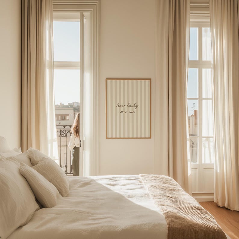 May include: A bedroom scene with a bed covered in white linens and pillows. A framed artwork with vertical stripes and the text "how lucky are we" hangs on the wall. Beige curtains frame a window and balcony.