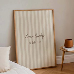 May include: A framed vertical print with a striped pattern in beige and off-white. The text "how lucky are we" is written in a cursive font. A small wooden side table with a brown ceramic pot and a book is next to the print.