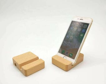 May include: A wooden phone stand with a slot for the phone to rest in. The stand is made of light-colored wood and has a simple, minimalist design. A white smartphone is displayed on the stand. The stand is designed to hold a phone upright.